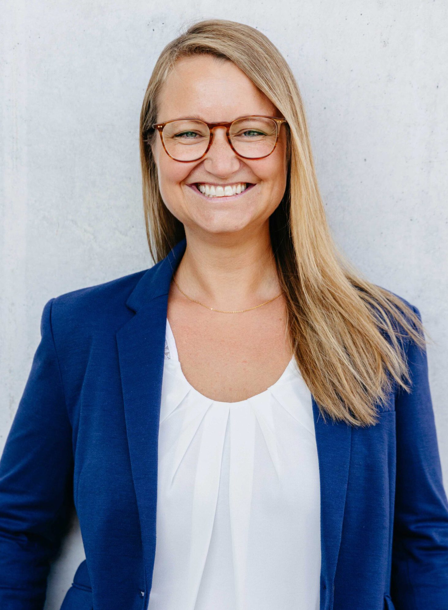 Lächelnde Businessfrau mit blauer Brille und blauem Blazer lehnt entspannt an einer weißen Betonwand.