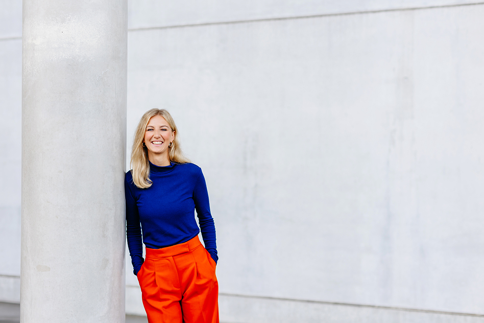 Blonde Frau in blauem Oberteil und orangefarbener Hose lehnt lächelnd an einer hellen Betonsäule vor moderner Architektur.