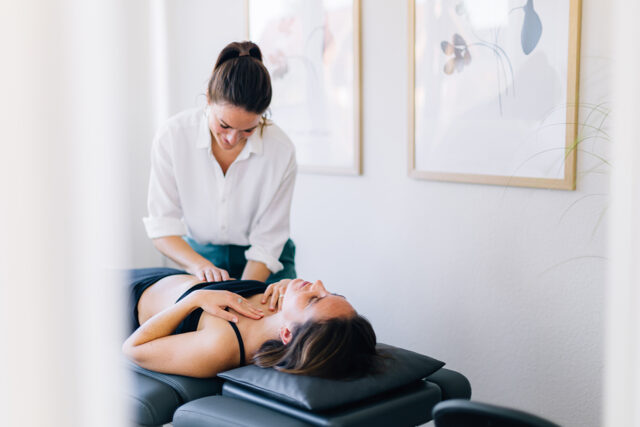 Physiotherapeutin mit zusammengebundenen Haaren und weißer Bluse behandelt eine liegende Patientin auf einer Therapieliege in einem hellen Praxisraum mit gerahmten Bildern an der Wand.