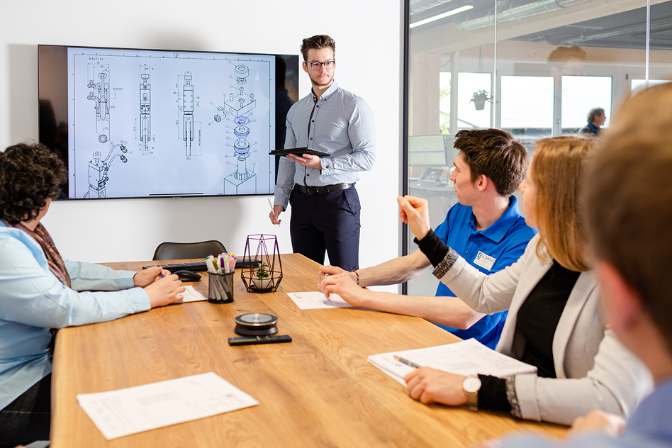 Mann steht vor einem Bildschirm mit technischen Zeichnungen und erklärt diese einem Team an einem Besprechungstisch.