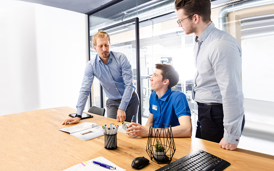 Drei Männer stehen und sitzen an einem Holztisch und besprechen gemeinsam Unterlagen in einem modernen Büro.