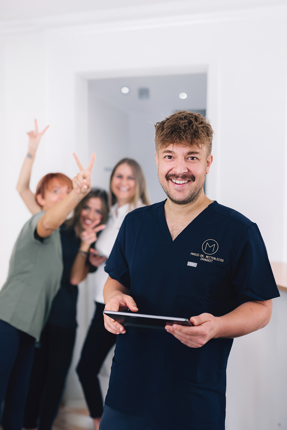 Mann in dunkler medizinischer Arbeitskleidung hält ein Tablet in den Händen und lächelt in die Kamera, während drei Frauen im Hintergrund in einem hellen Praxisraum gestikulieren.