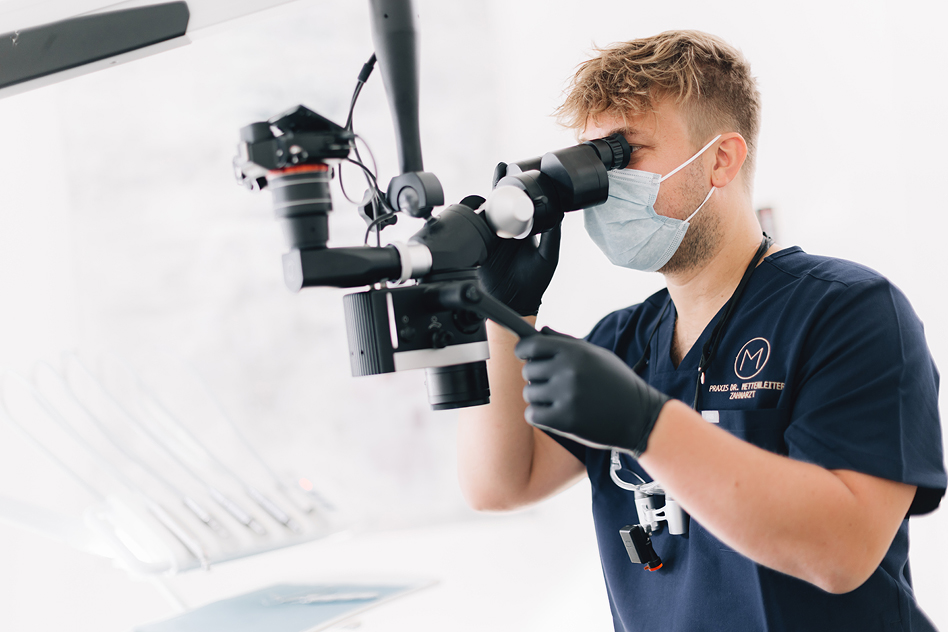 Mann in dunkler medizinischer Arbeitskleidung trägt eine Schutzmaske und schwarze Handschuhe und blickt durch ein Behandlungsmikroskop in einem hellen Praxisraum.