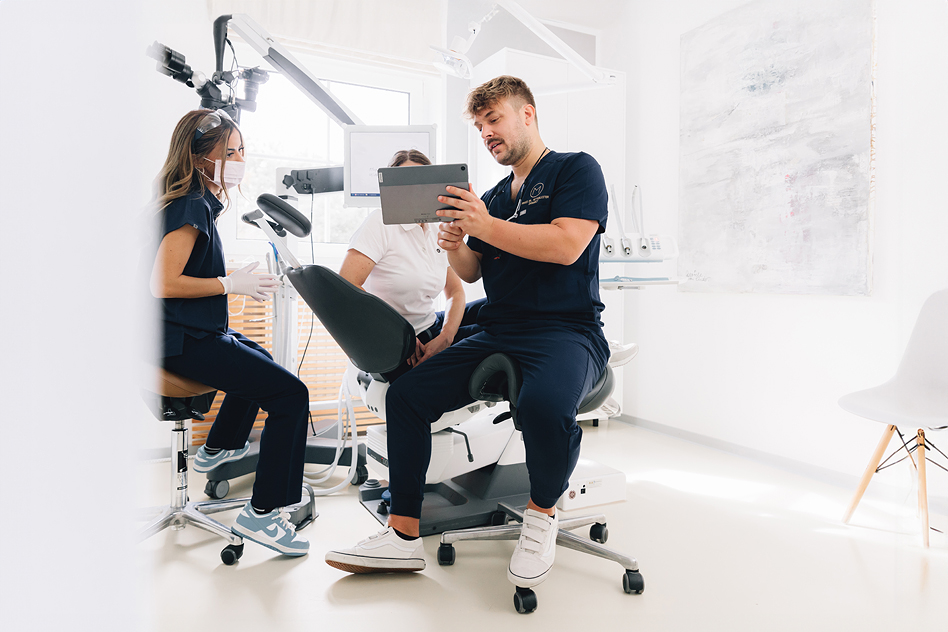 Zahnarzt sitzt in einer modernen Zahnarztpraxis auf einem Behandlungsstuhl und zeigt zwei Mitarbeiterinnen auf einem Tablet etwas; eine Assistenz trägt Mundschutz und Handschuhe, im Hintergrund steht ein Zahnarztstuhl mit Behandlungslampe in einem hellen Praxisraum.